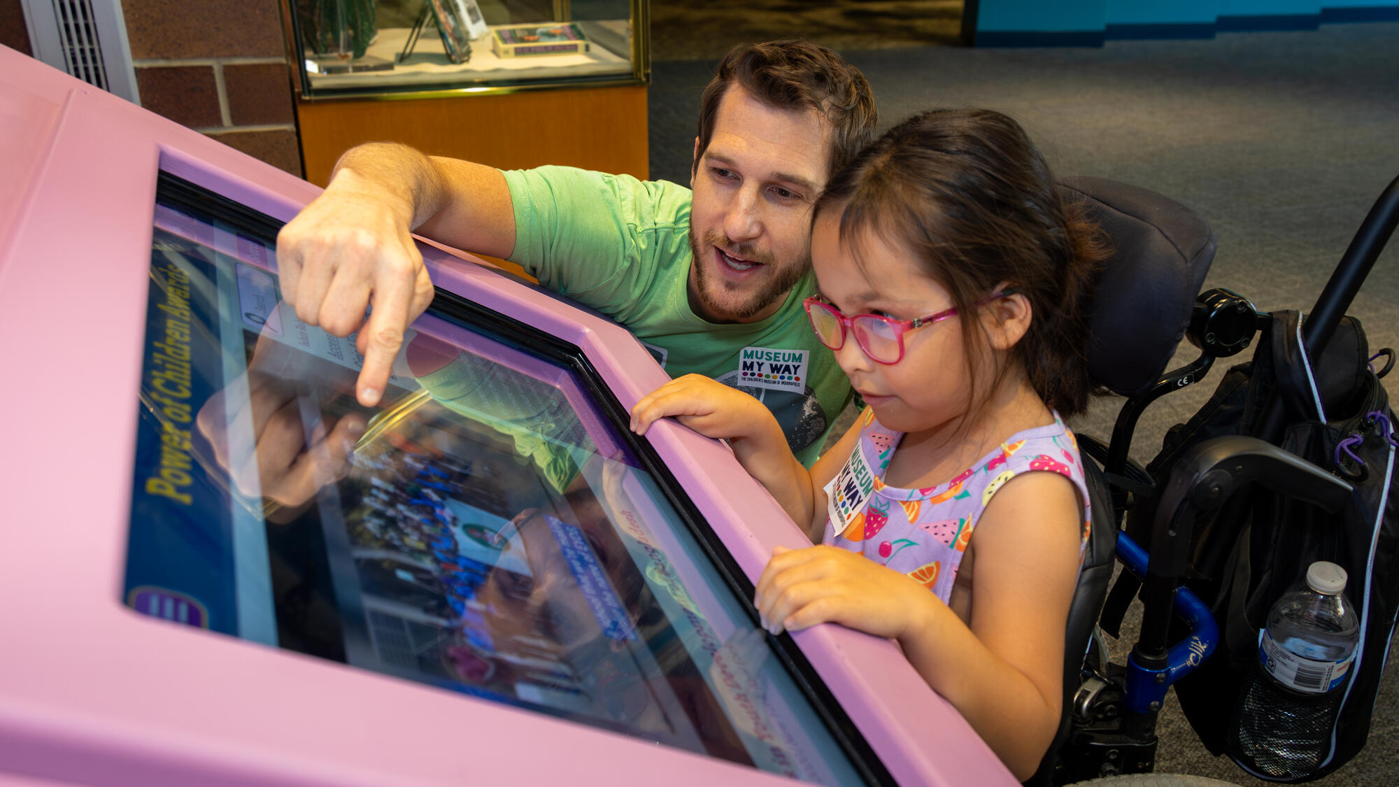 A child using a wheelchair and an adult view a touchscreen outside the Power of Children exhibit. The adult is crouching down to the child’s level and points to the screen.